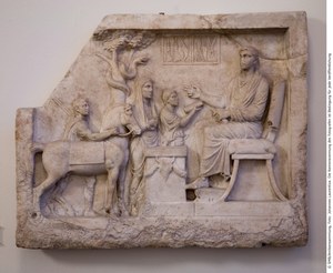 image showing a section of marble carved into a relief, depicting a seated male figure, three standing figures, a horse, a snake in a tree, and a box of surgical instruments