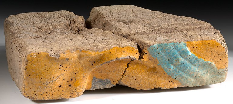 Two parts of a rectangular broken brick are placed together along the fault. The face of the brick is molded and painted yellow with a stripe of light blue on the right. The top of the brick, visible in the photograph, is unpainted gray-brown clay. The brick is photographed against a black background.