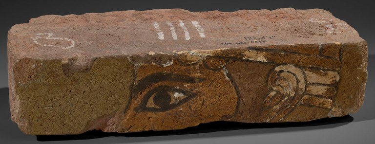 A rectangular brick with a part of a god's face on the face of the brick. The eye and ear are prominent. The brick is light brown with hints of black, white, and light green pigment.