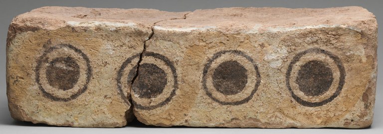 A brick broken into two parts. On the edge of the brick, there are 4 designs similar to eye motifs with a large dot surrounded by another circle in black pigment. The brick is otherwise unpainted brownish clay.