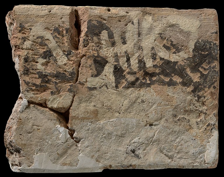 Parts of a broken brick in three pieces are placed together along the faults to create the shape of a rough rectangle. The brick is painted in a faded and chipped black pigment, with white handwritten markings along the top of the brick's painted face. The brick is photographed against a black background.
