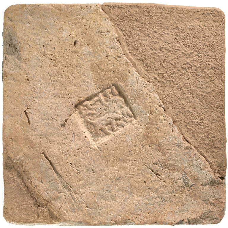 A square brick made of light brown clay, stamped at center with an alphabetic inscription and a lion. The brick has rounded edges and has two major cracks on either side of the inscription.