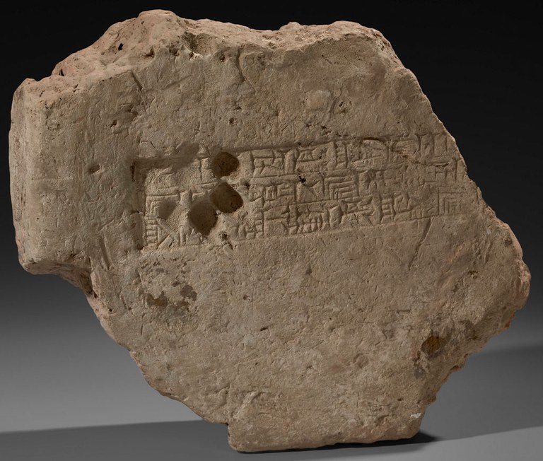 Grayish-brown clay brick fragment with chipped edges. The brick is stamped with three rows of cuneiform text in a rectangular block in the center, and also impressed with a dog's paw print on the left side of the text.