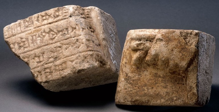 Photograph with two roughly cube-shaped pieces of fragmentary bricks. One piece has cuneiform inscriptions with straight lines between the rows of symbols and the other shows the top of a closed hand in relief. Both bricks are a brownish color.