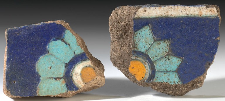 Two corner pieces of a broken tile with rosette motifs on it. The tile is glazed and of siliceous material. The colors on the tile are yellow, navy blue, turquoise and white.