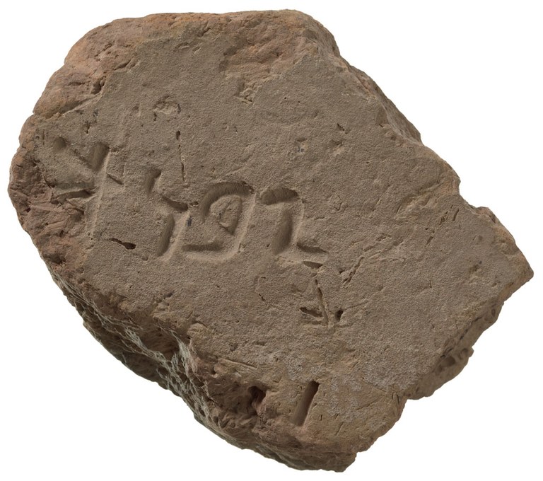 A dark gray brick fragment, roughly rectangular and chipped at its edges, with an alphabetic inscription on it, center left.