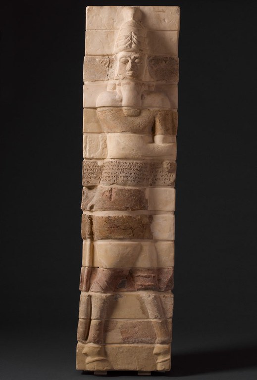 Rectangular pillar with bricks in molded relief showing a man with a long beard and a tall hat, as well as bull-like hooves for legs. The whole figure is divided into 14 stacked horizontal bricks and the brick at the mid section has a cuneiform inscription carved into it. The bricks are shades of brown with some variety of shade.