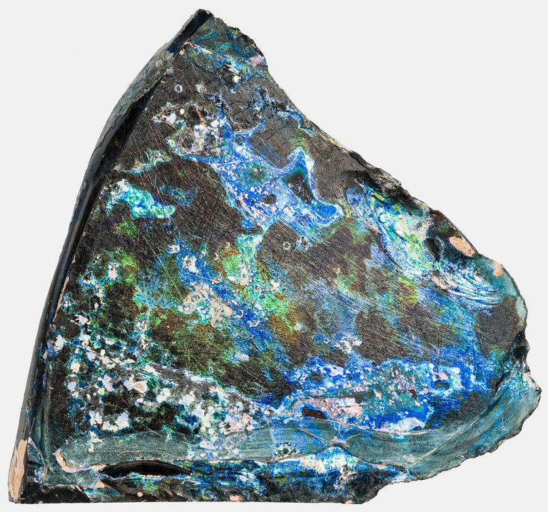 A piece of a cullet, made entirely out of glass. The fragment is irregularly shaped, multi-colored with green, blue, navy blue, white, black and a light pink hues mixed within.