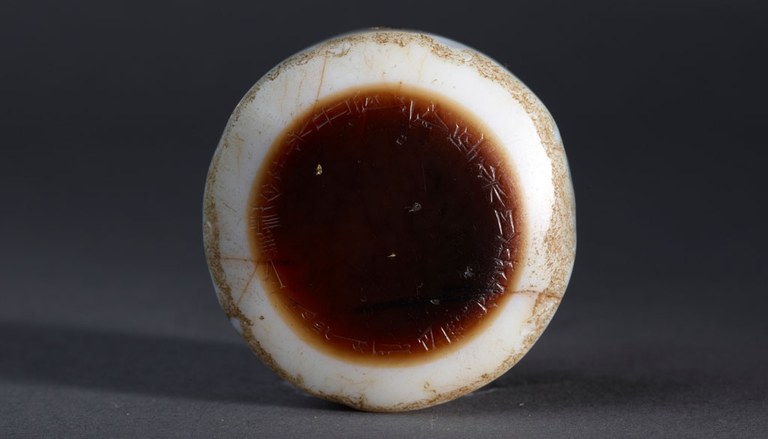 An image of an eyestone made of agate with cuneiform inscriptions on it in a circular pattern. The center of the stone is a deep orange-brown color encircled by a white border. The inscription is in the brown portion of the stone, and the outer white band shows signs of wear at the edges.