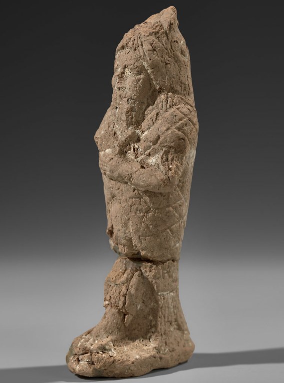 Molded and hand worked unbaked figurine of a standing human figure wearing a fish skin. The figure is light brown with a rough surface and shows signs of wear.