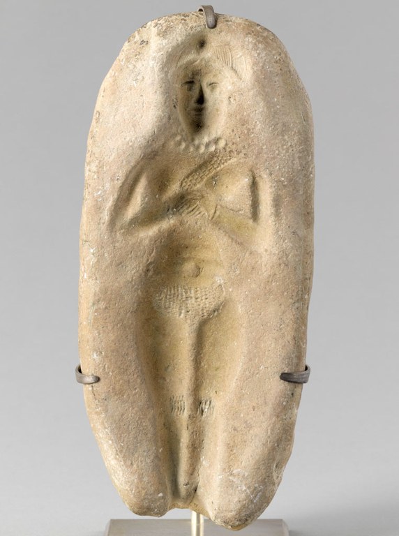 Mold showing impression of a standing female figure with hands clasped in front of her chest and holding something rectangular. She wears a bead necklaces and possibly a hat or headdress. The mold is oval-shaped and the figure's feet are absent.