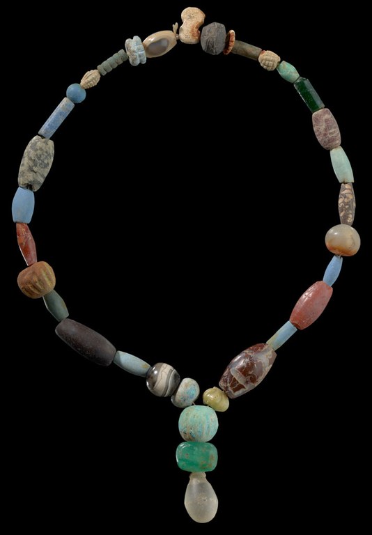 Green, blue, purple, white, yellow, and a few stripped black-and-white stones, in different rounded shapes and sizes, strung together forming a necklace. There is an additional stone pendant with light blue, green, and semi-translucent white stones. The necklace is photographed against a black background.