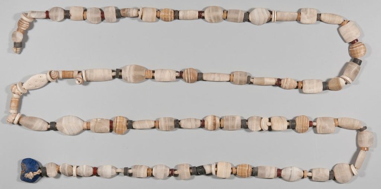 A necklace made of stones, shells, and other materials with holes in them is laid out on a light background in serpentine fashion. The stones are mostly light-colored, some with white stripes. The shapes vary, from small disks to oblong and oval shapes. One end is finished with an unsymmetrical, large blue stone.