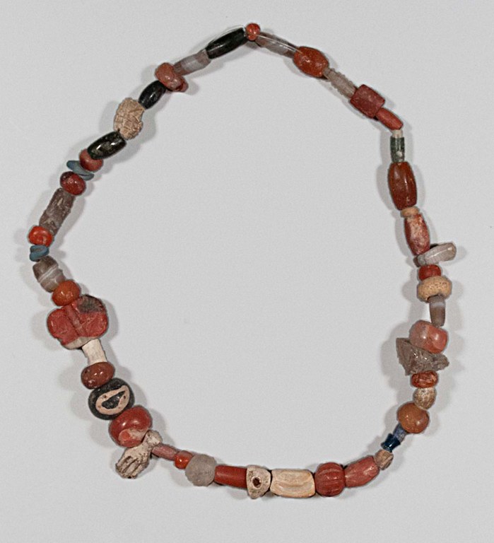 A reconstructed necklace made differently shaped beads of stone, agate, rock crystal, shell, and vitreous materials. The bead colors are a mixture of browns, red, dark red, ivory, orange, dark grey, and black.
