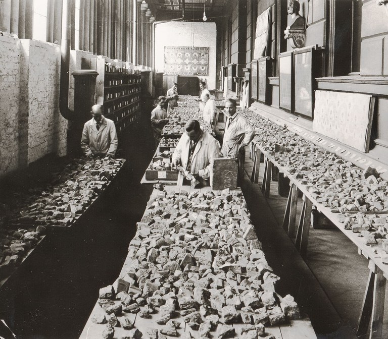 Black and white photographs of a brick fragment in a laboratory-type setting. The fragments of bricks are laid out on long tables, arranged and sorted by a few men.