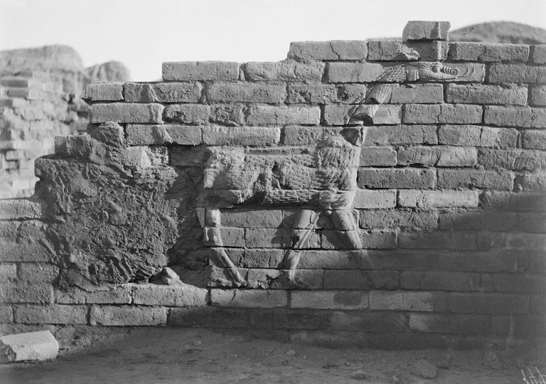Black and white photograph of an unglazed dragon in raised relief brick, part of a fragmentary wall. The dragon, with an elongated neck and the legs of a bull, is facing left and his hind leg and rump are eroded.