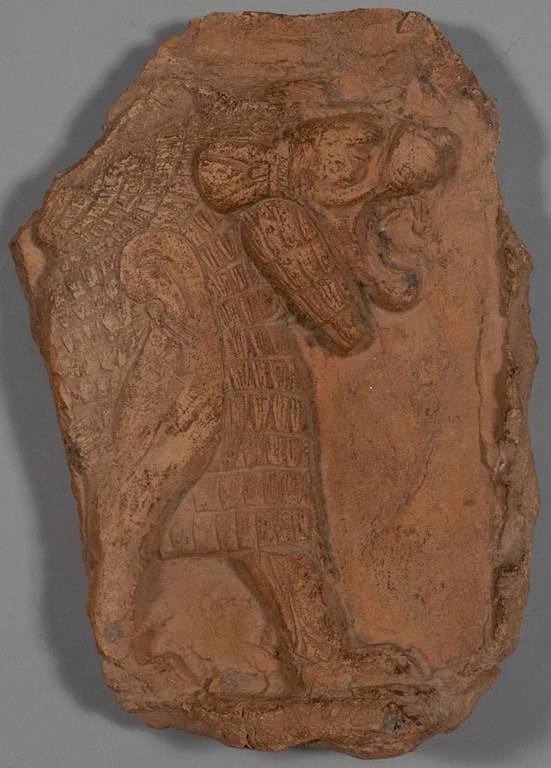 A clay tablet with chips missing from the corners and edges. On the face of the tablet is a molded, roaring lion in profile.
