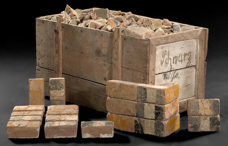 A wooden crate with an open top filled with brick fragments and the word "Schmarz" written by hand in faded black paint on one side of the box. 16 bricks with faded and cracked yellow, black, and white pigment bands are arranged in front of the crate. The crate and bricks are photographed in front of a back background.