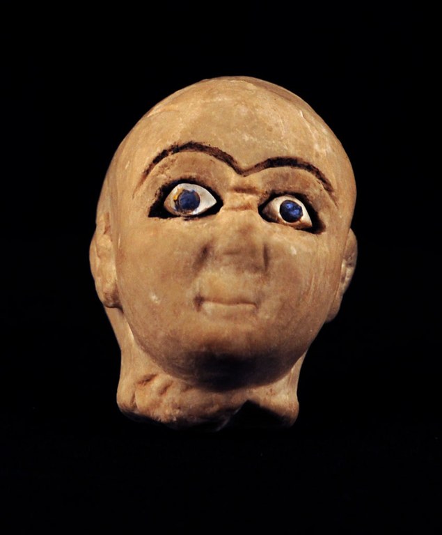 Head of a statue of a worshipper or priest. The composite statue is one without any hair on the head. It has wide eyes, orange-hued skin, and a broken nose.