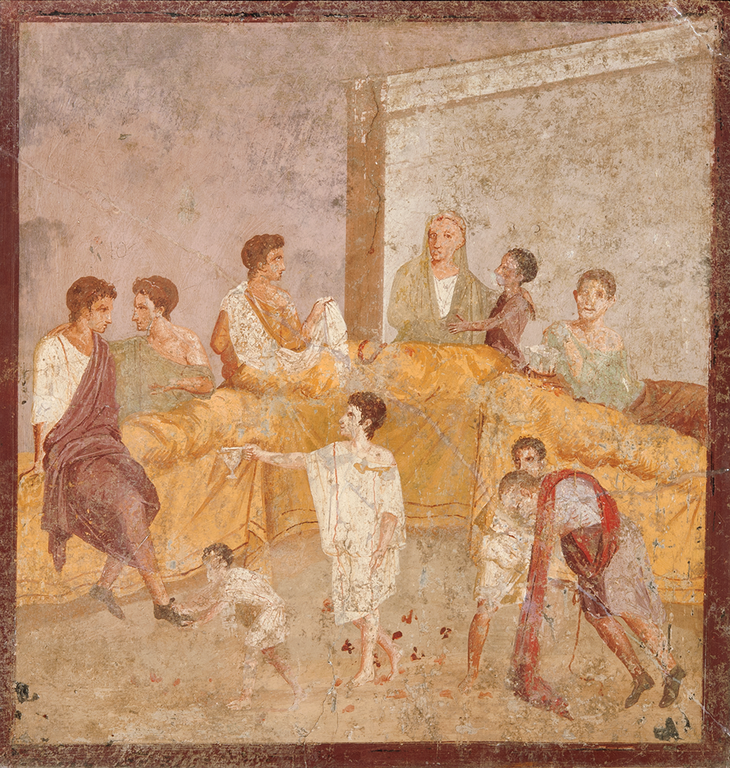 Four men recline on long beds draped in cloth for a banquet or symposium. Another man is seated on the front edge of the table. He speaks to the figure at his left, and scratched above his head is the Greek word Scio (I know). He looks at a smaller male, likely enslaved, in front of the beds, who extends a silver cup to him. An even smaller male, possibly a child, appears to help the man sitting on the table with his shoe. At the right a third small figure supports a reveler who appears to be incapacitated; the word Bibo (I drink) is scratched above him. At the center of the reclining guests is a man whose white hair is covered with a cloth. A small, dark-skinned boy is seated on his lap. Scratched above the man’s head is Valetis (Be Well), and above the boy, Isisa, possibly his name.