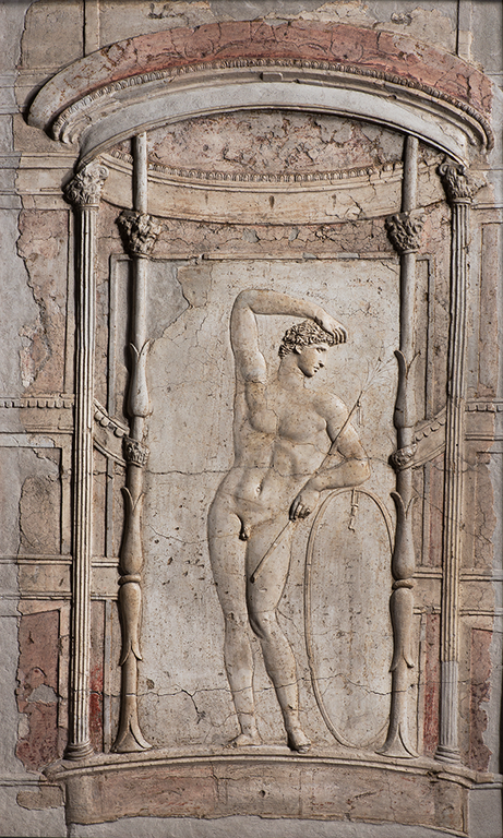 At the center is the stucco relief of a nude male within a colonnaded architectural frame. He supports his weight on his right leg. He bends his right arm to touch the top of his head. In his left hand he holds a long rod. There is no pigment on this work and the overall color of the image is reddish-gray.