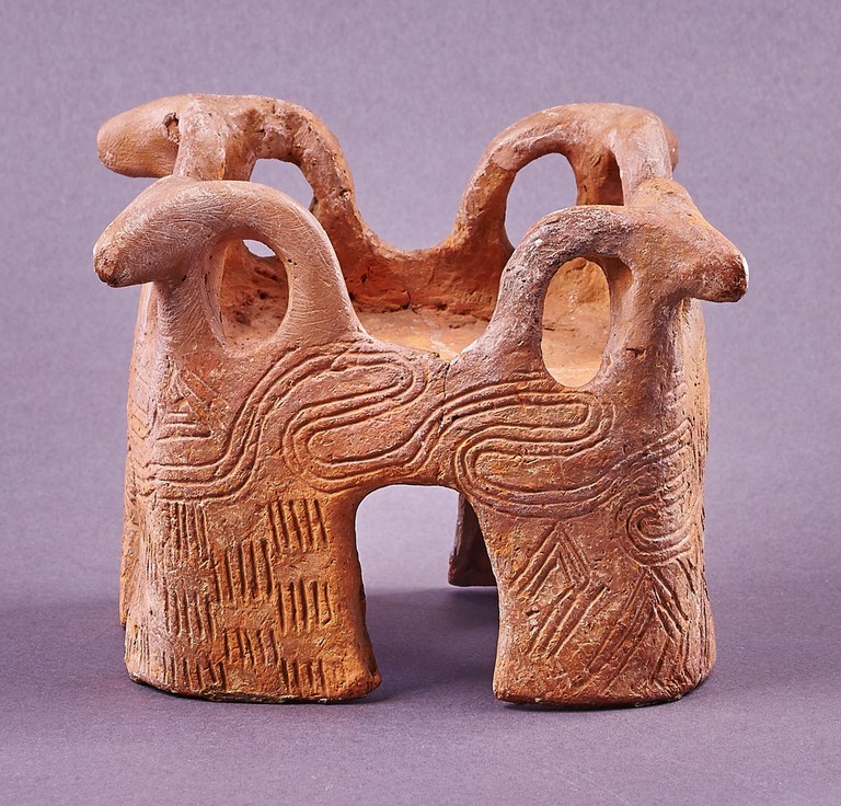 A table-like form with four wide legs. The vertical surfaces are incised with spiral and meander patterns. Each of the legs is topped with a stylized ram's head.
