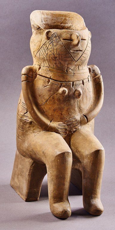 A female figure seated on a chair. Her face is decorated with incised lines within a broad M shape, above which incised lines indicate eyes and a mouth, with clay projecting for the nose and ears. The back of the head extends upward. Her hands are clasped in her lap.