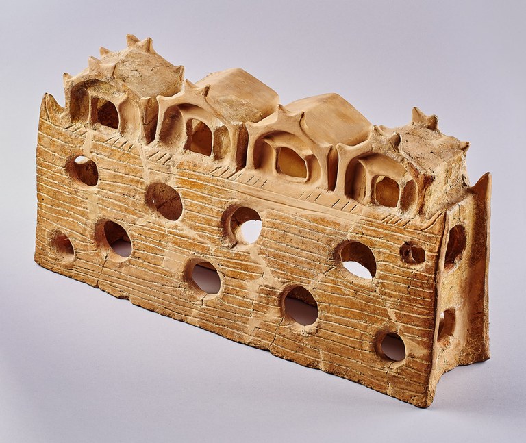 A long, hollow rectangular form resembling a building. The base is perforated with large round holes and incised with horizontal lines. At the top, four gables with openings have horn-like extensions at the front and back.