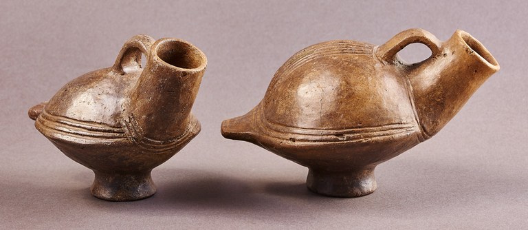 Each vessel takes the form of a stylized bird with a rounded body, a tail, and a long spout that resembles a neck. The top of each spout is attached to the body with a handle. The foot on each vessel suggests a bird's legs.