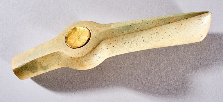 A long rectangular piece of stone, flat at one end and tapered at the other. About midway along its length, the form extends to a circle and is inset with a flat piece of gold perforated with small dots.