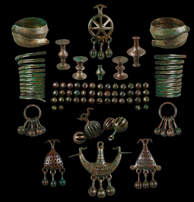 Two bracelets with tapering ends. Two spiral bracelets. One rattle consists of a spoked wheel and a handle. Three slotted cases with beads hang from small rings at the bottom of the wheel. Two of the rattles consist of a single ring from which hang round slotted cases with beads. Two triangular pendants and one crescent-shaped pendant are decorated with triangular perforations and have hanging beads similar to those on the rattles. Four of the five belt fittings consist of wide flat bases joined by circular forms at the midsection. One of the belt fittings has narrow ends, each of which tapers outward to the midsection. The wand is composed of four round slotted cases, one of which is topped by a birdlike form. Forty-three loose beads.
