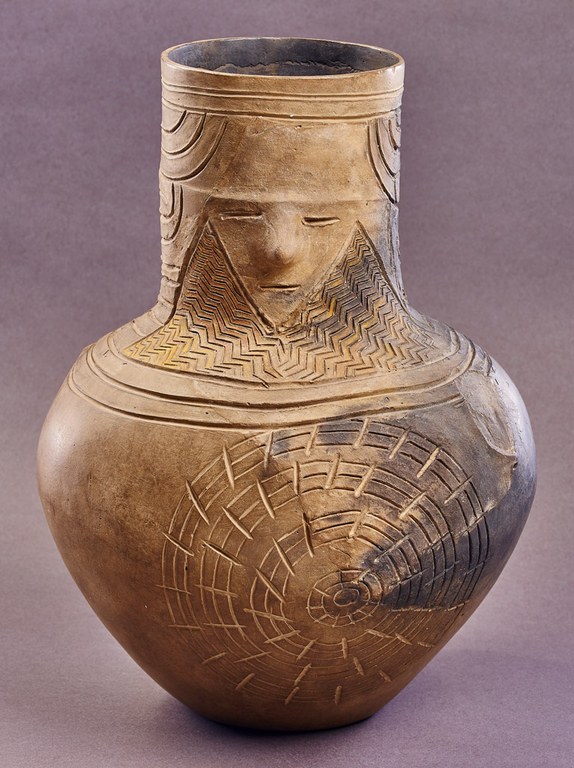 A light brown pot incised at the neck with two horizontal slits for eyes, a projection for the nose, and one horizontal slit for a mouth. The neck and shoulder are incised with linear patterns. The body is incised with a series of concentric circles intersected by short lines.