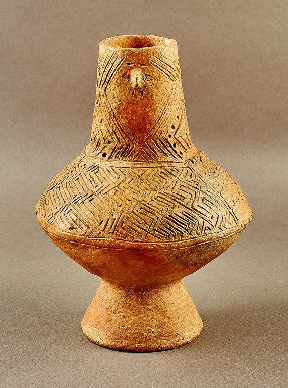 A sand-colored pot incised at the tall neck and the shoulders with angular patterns. At the center of the neck, clay projects from the surface to depict a nose, and incised lines suggest eyes and a philtrum. The body is composed of two convex forms.