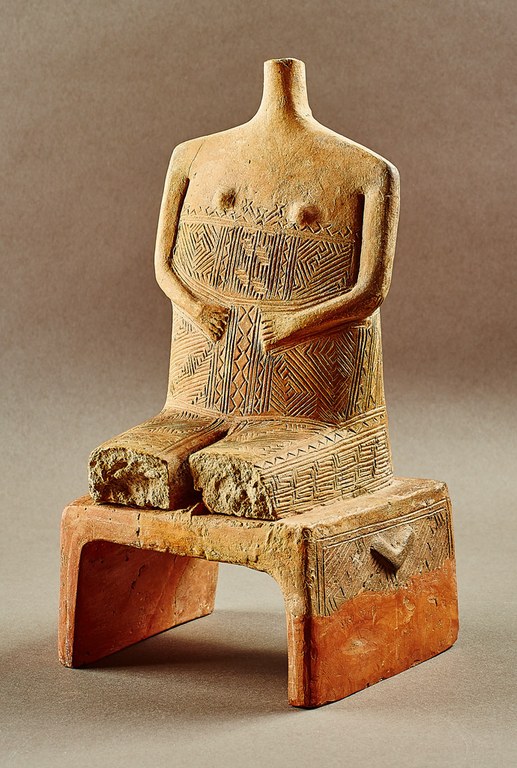 A headless female figure seated on a stool. The torso and legs are decorated with incised linear patterns. The legs have been broken just above the knees so that only the thighs remain.