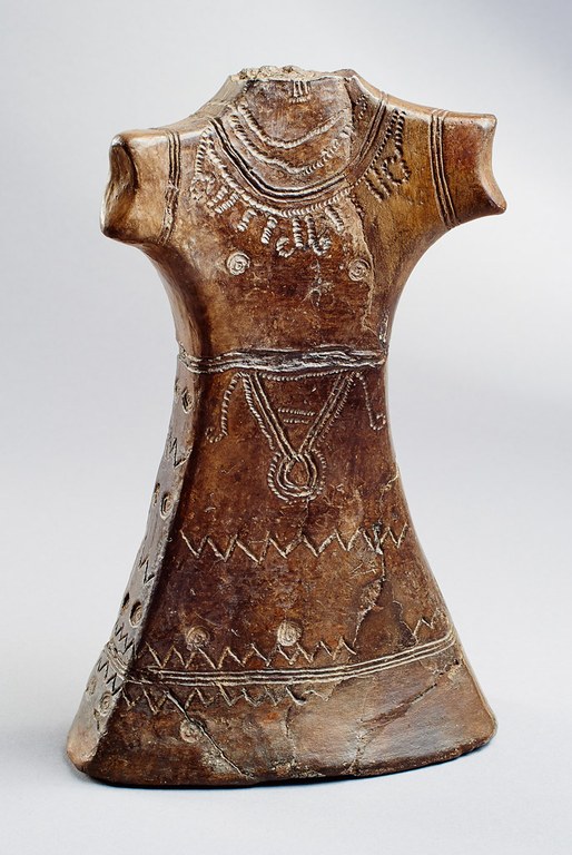 A figure wearing a women's dress incised to suggest embroidery as well as jewelry. The head and arms are missing. The bottom of the figure ends in a flat base suggesting the hem of the dress.