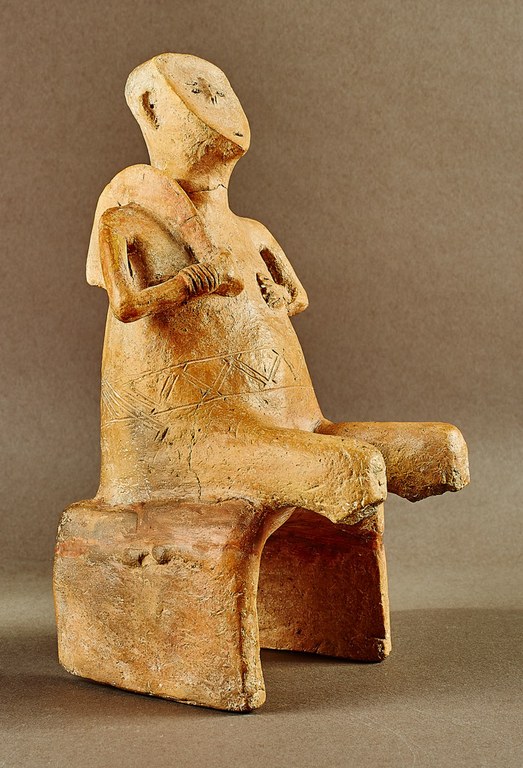 A sand-colored male figure seated on a chair. The face is flat with incisions for the eyes and mouth, and a projection for the nose. An incised belt encircles his waist, and he holds a long curved tool over his proper-right shoulder. His wrists are incised to indicate bracelets. The legs are broken below the knees.