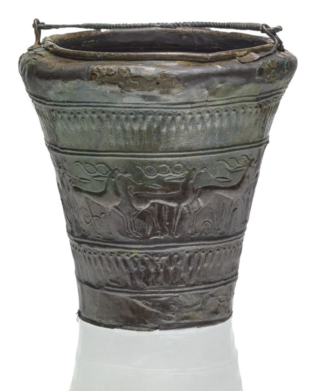 A bucket-shaped vessel decorated with a procession of stags and abstract ornaments. The handle is long piece of spiralized metal.