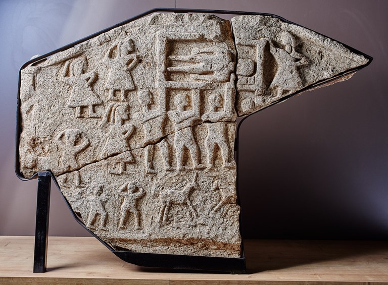 Fragments of a large stone depicting males and females and one male in an open, box-like grave.
