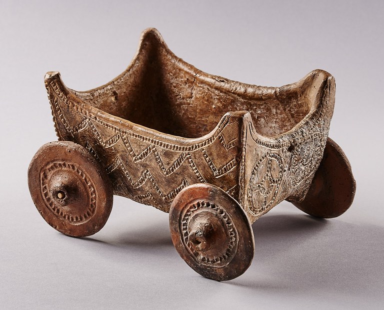 An open boxlike form with peaks at each corner and four flat circular wheels. The wagon is decorated with incised geometric designs.