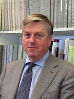Photograph of Alexander Jones, wearing a suit and tie and standing in front of full bookshelves.