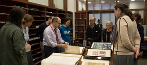 A group of people stand around a table full of large, open books, apparently having a discussion.