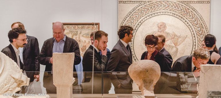 A group of people examine ancient artifacts in a large gallery display. Large mosaics in frames are mounted on the walls behind them.