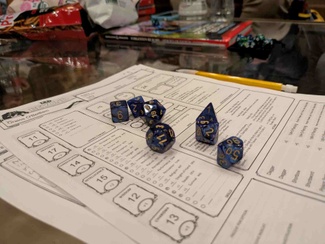 A collection of dark blue polyhedral dice on top of a Dungeons & Dragons character sheet (for О̄raios O'Bedlam, a tiefling rogue). Out of focus in the background are additional dice, a pencil, a Dungeons and Dragons player handbook, and a bag of pretzels.