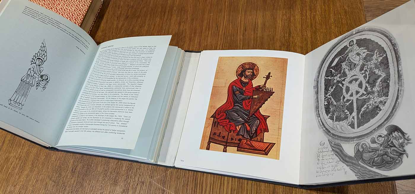 An open book in an unusual binding: it has two separate text blocks that are open side-by-side. The left side shows an image of an angelic being holding the head of a, saint with English-language text (too small to be legible in this image); the right side shows a color image of a saint playing a musical instrument and a black-and-white image of a heavenly scene of Christ and several angels.
