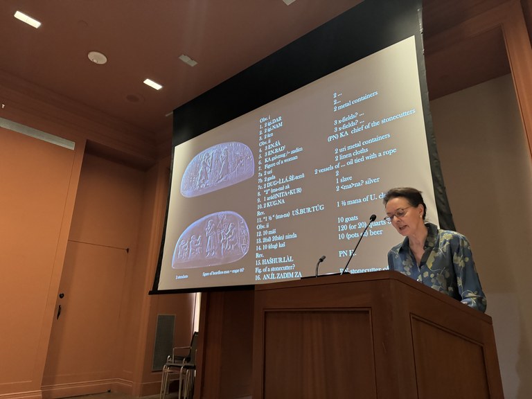 A woman at a podium in a lecture hall presenting; behind her a screen with two images of an ancient inscribed object on the left and transliterated and translated text on the right.