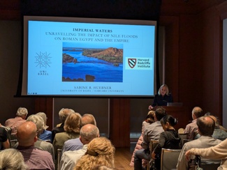 Prof. Sabine Huebner lecturing in front of a seated audience with the opening slide of her presentation, "Imperial Waters," behind her