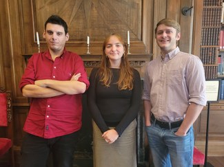 Three individuals stand side-by-side in front of a wood-paneled wall with a fireplace and bookshelves partly visible behind them.