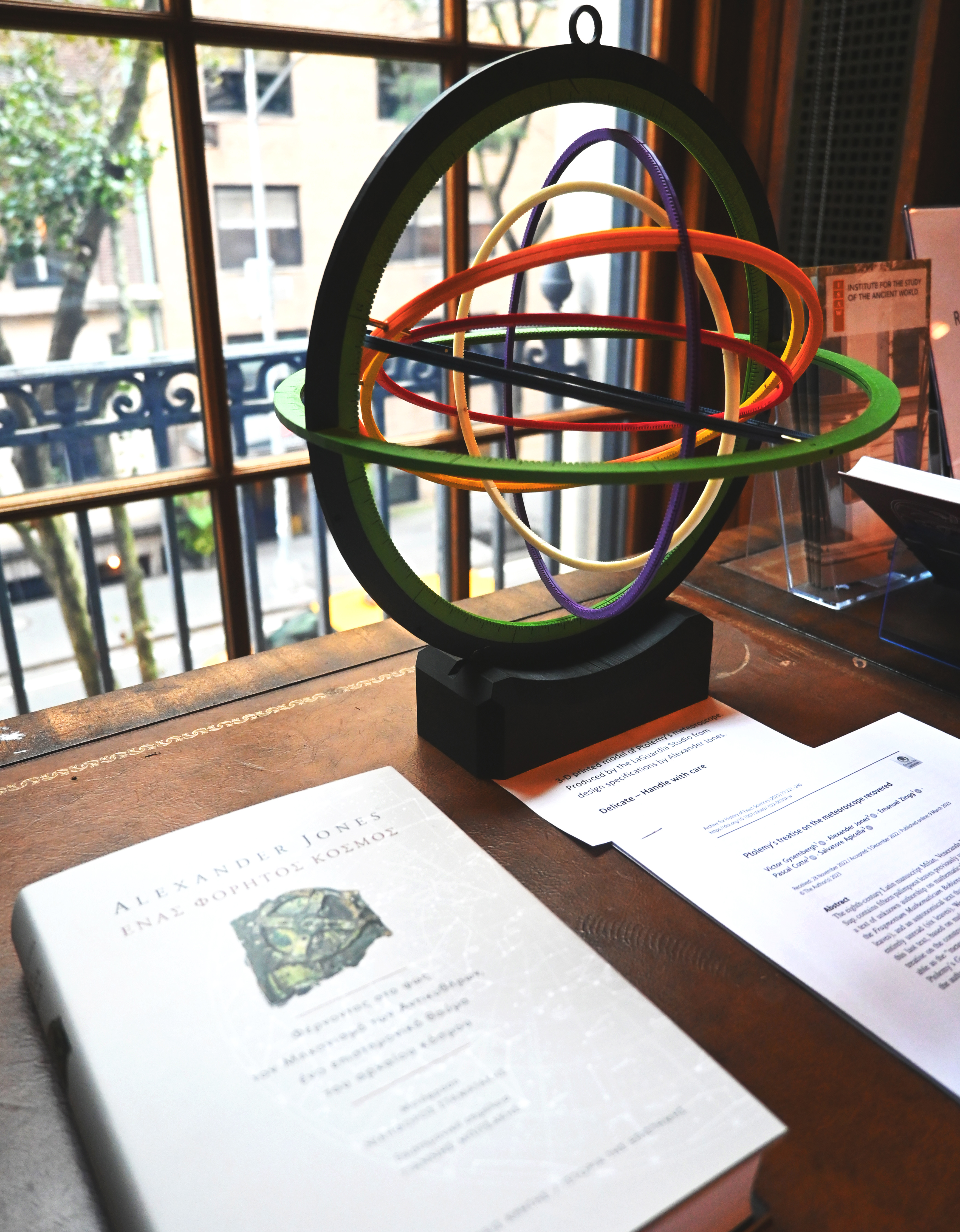 Book titled A Portable Cosmos written by Alexander Jones translated into Greek and displayed on a table next to a model of an ancient meteoroscope