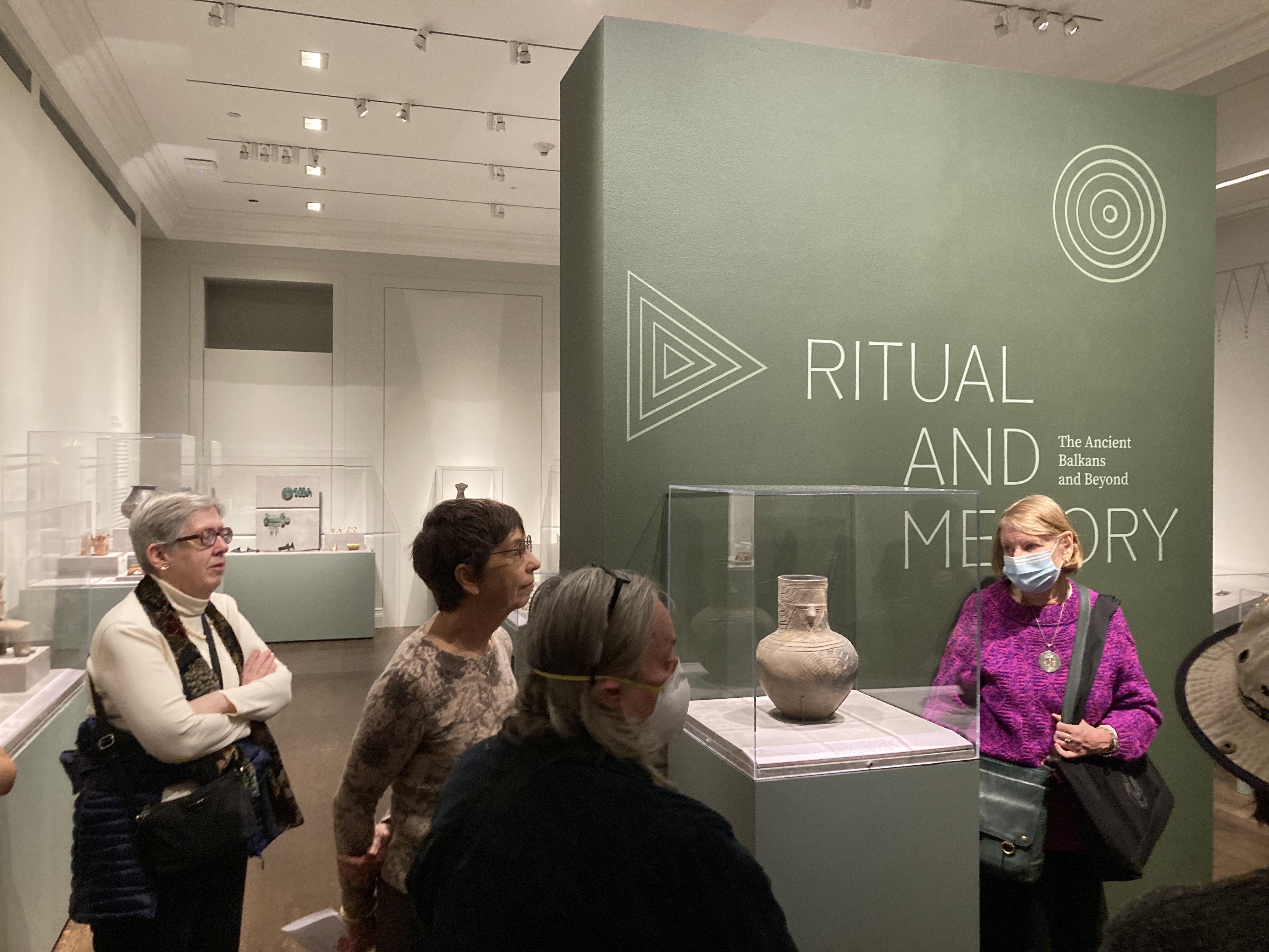 four people looking at a ceramic vessel with a human face inside a glass case in front of the exhibition banner labeled “‘Ritual and Memory: The Ancient Balkans and Beyond