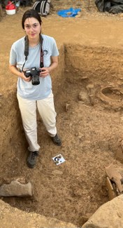 A woman wihth a camera on a strap around her neck stands in a rectangular, excavated area. She looks up at the camera.